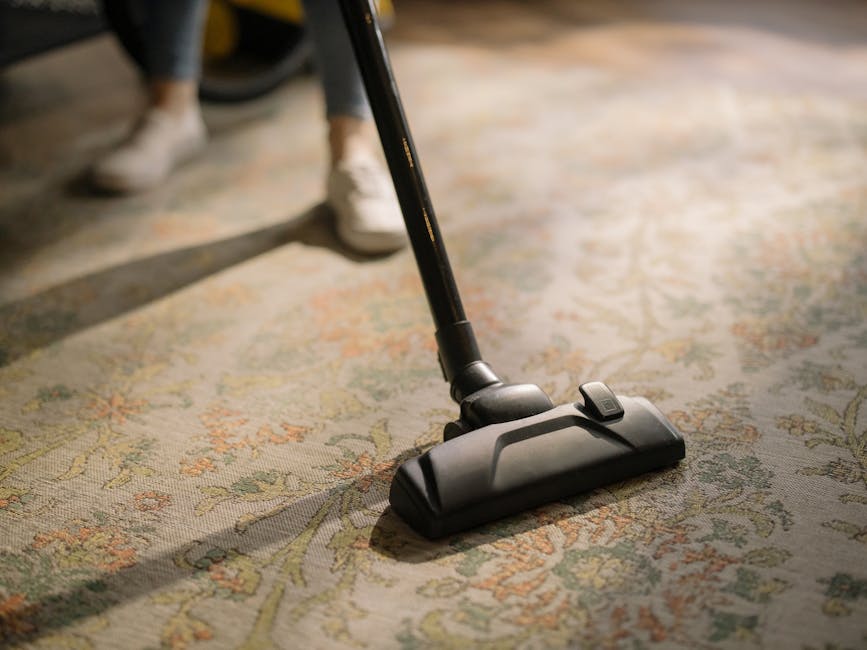 A person using a black vacuum cleaner with a slim nozzle attachment to clean a patterned, beige and pastel-colored carpet in a well-lit room. The person's legs are visible, wearing dark pants and ligh