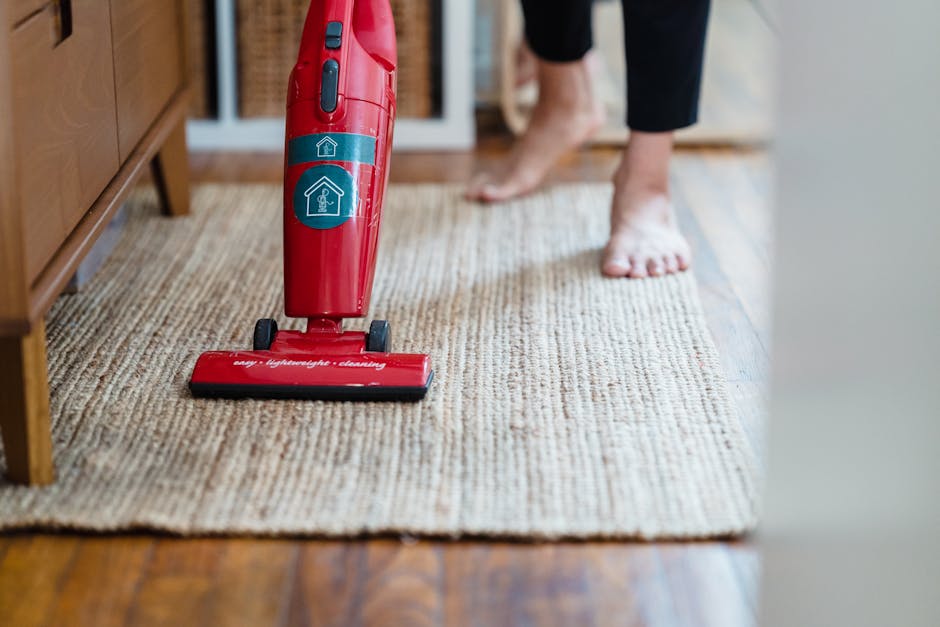 A person using a black vacuum cleaner with a slim nozzle attachment to clean a patterned, beige and pastel-colored carpet in a well-lit room. The person's legs are visible, wearing dark pants and ligh