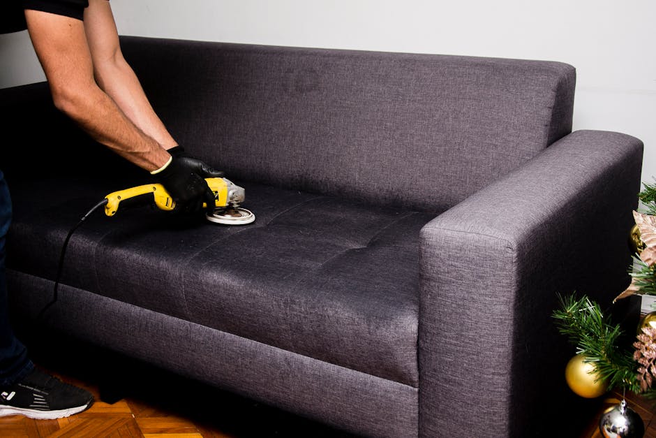 A close-up of a person's hand holding a handheld upholstery cleaning tool connected to a vacuum hose, performing deep cleaning on a beige fabric car seat or sofa with a textured, patterned surface. Th