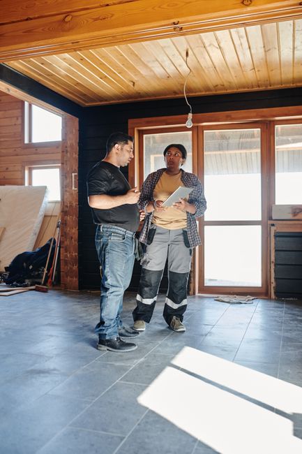 Two construction workers, a man and a woman, stand inside an unfinished room with a wooden ceiling and large windows, discussing plans while holding documents. The room features a tiled floor and part